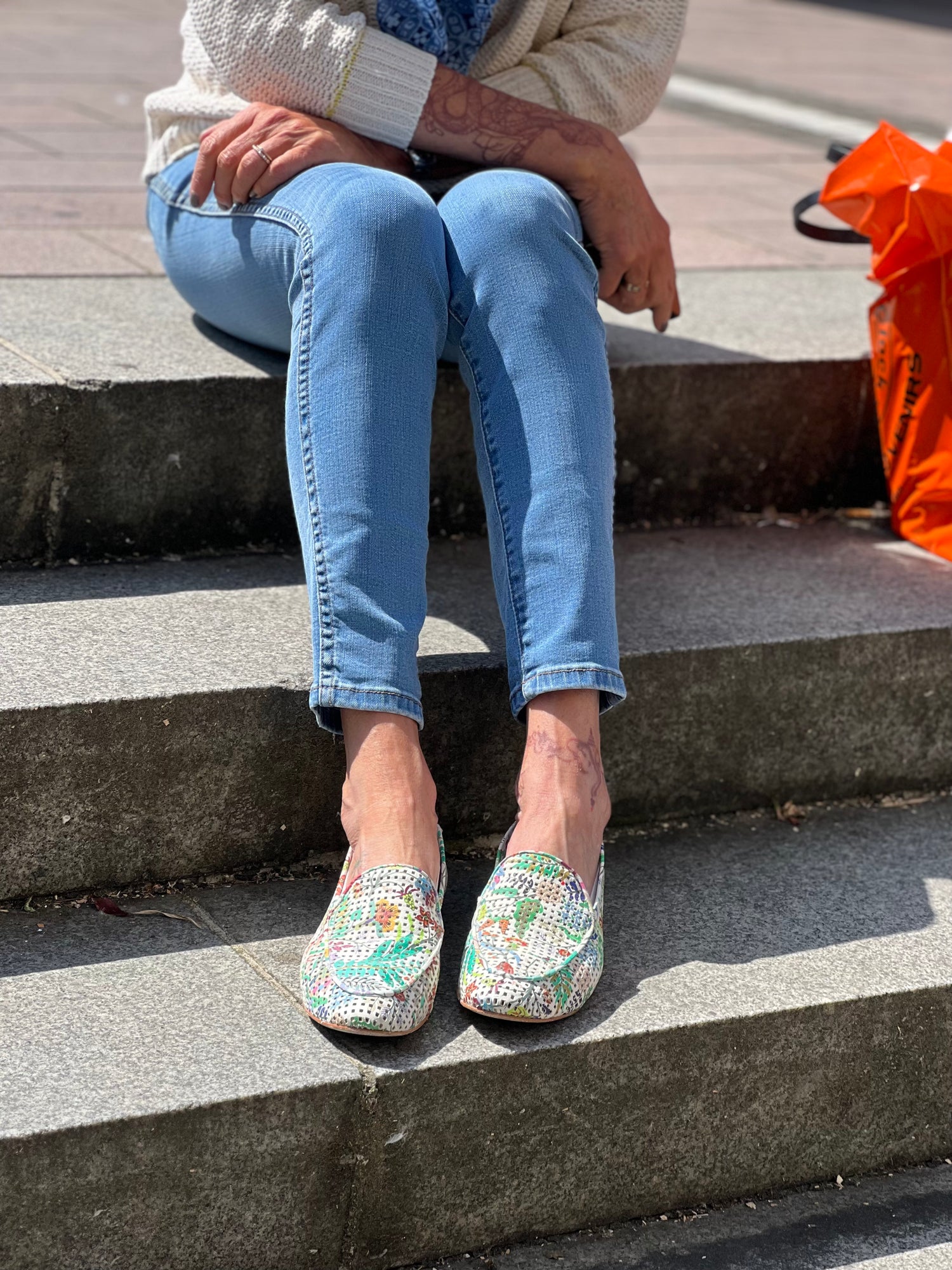 Person sitting on stairs wearing light blue jeans, a white sweater, and floral slip-on shoes with a blue scarf and orange bag beside them